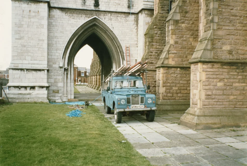 Photograph of a blue Land Rover loaded with ladders, parked on the paving beneath St Walburge's arched west porch, coiled ropes lying on the grass beside it. The pointed archway frames a further view of the covered walkway and houses beyond.