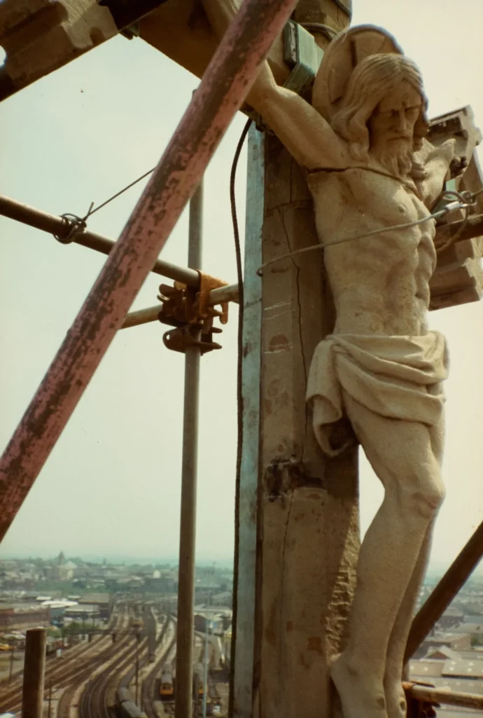 Photograph taken close beside the crucifix of St Walburge's Calvary, secured against its cross with a temporary wire during restoration, scaffolding poles crossing the frame. Preston's railway lines and rooftops visible in the distance.