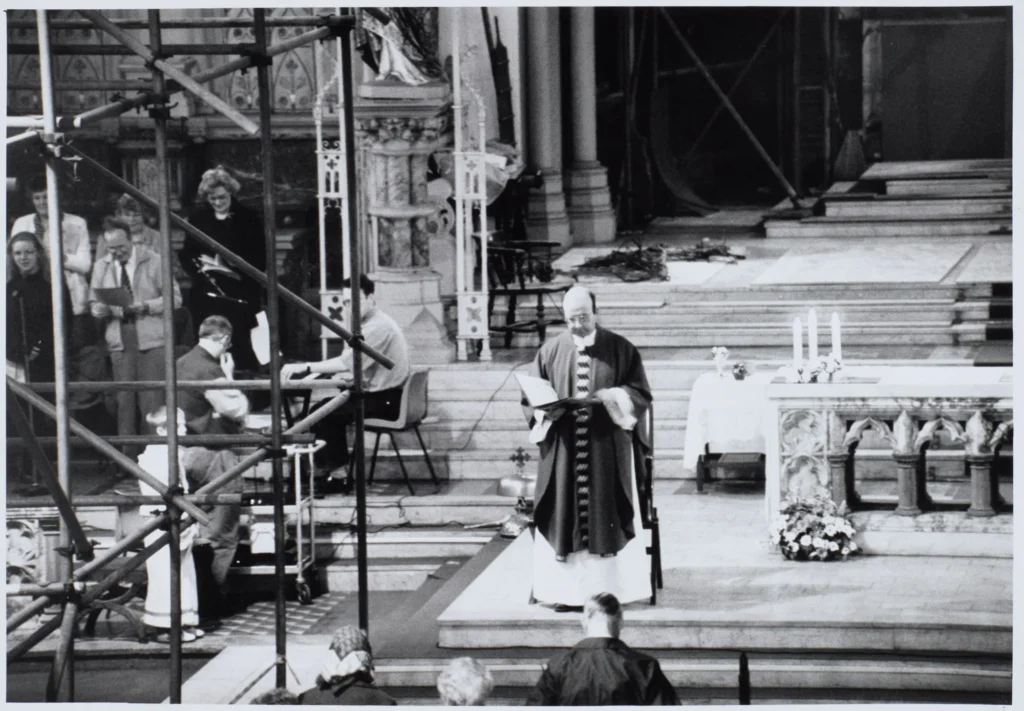 Photograph taken during at St Walburge's, with scaffolding crossing the sanctuary. A priest reads at the marble altar, candles and flowers behind the altar rails, while singers and an organist take part from the left.