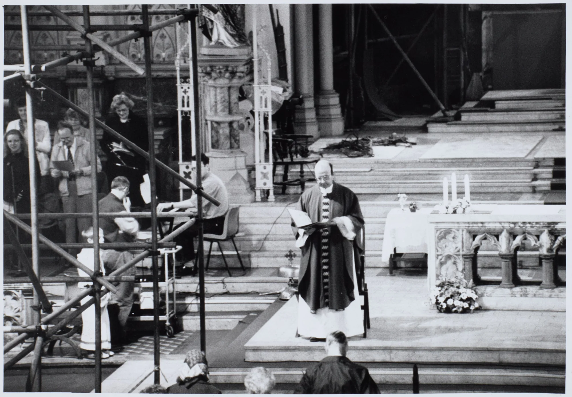 Photograph taken during at St Walburge's, with scaffolding crossing the sanctuary. A priest reads at the marble altar, candles and flowers behind the altar rails, while singers and an organist take part from the left.
