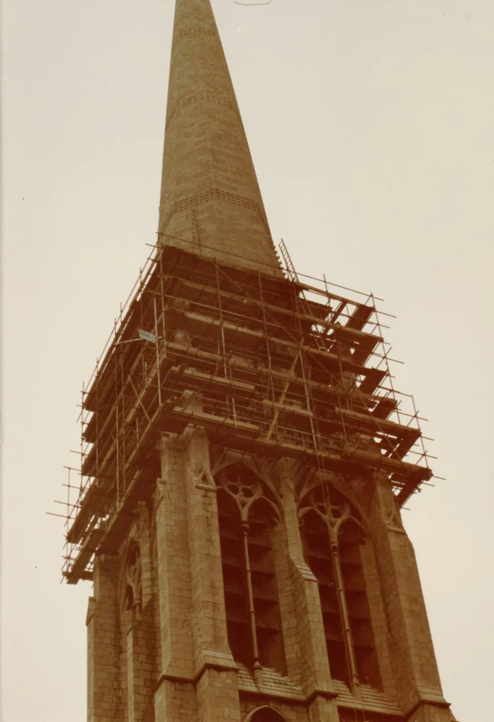 Photograph looking up at St Walburge's spire, scaffolding encircling the tower below the base of the spire while the stonework tapers unobstructed above. Gothic belfry windows are visible in the tower beneath.