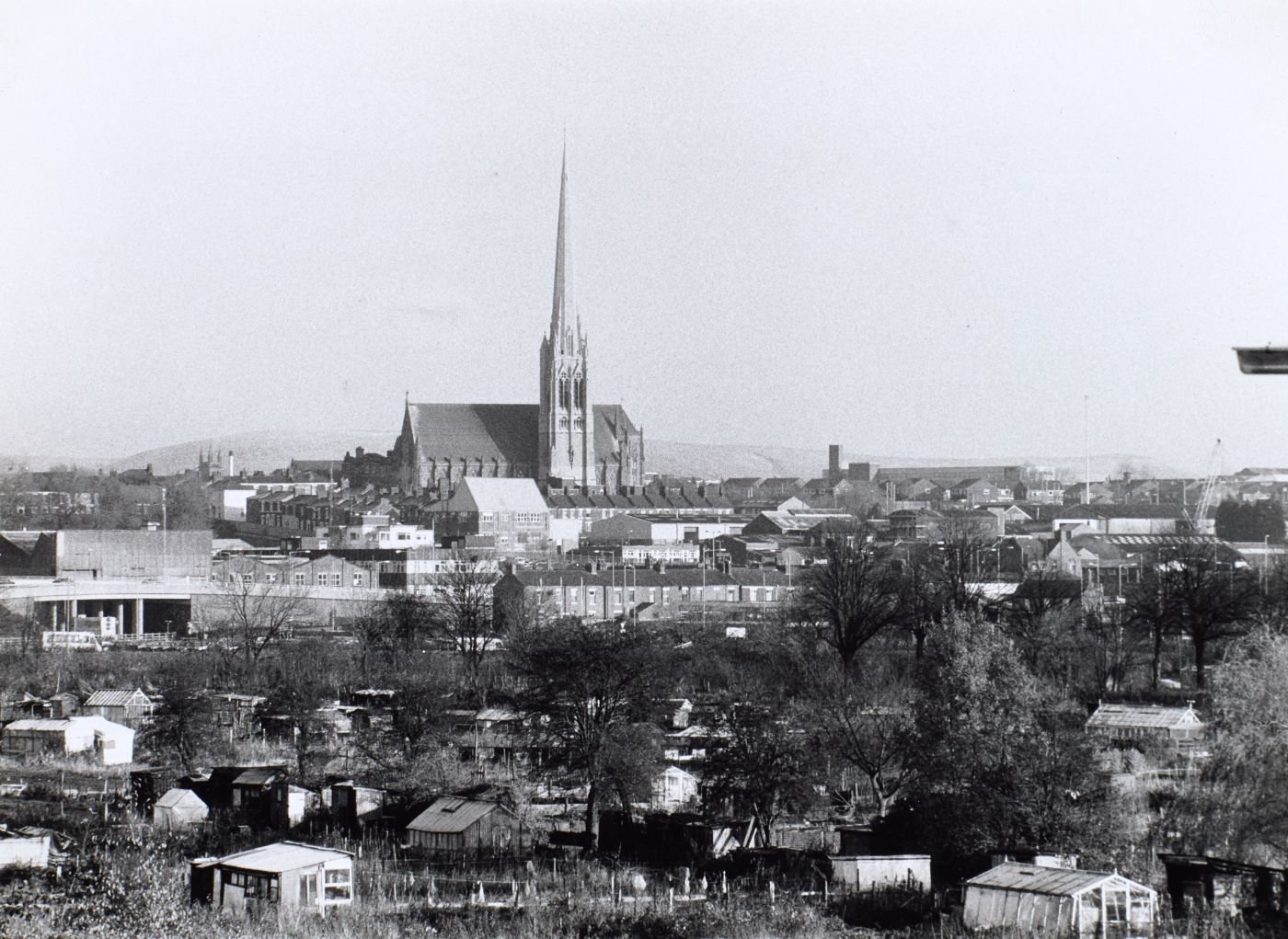 Photograph of St Walburge's spire rising above the town, allotments and greenhouses filling the foreground, a road flyover visible to the left.