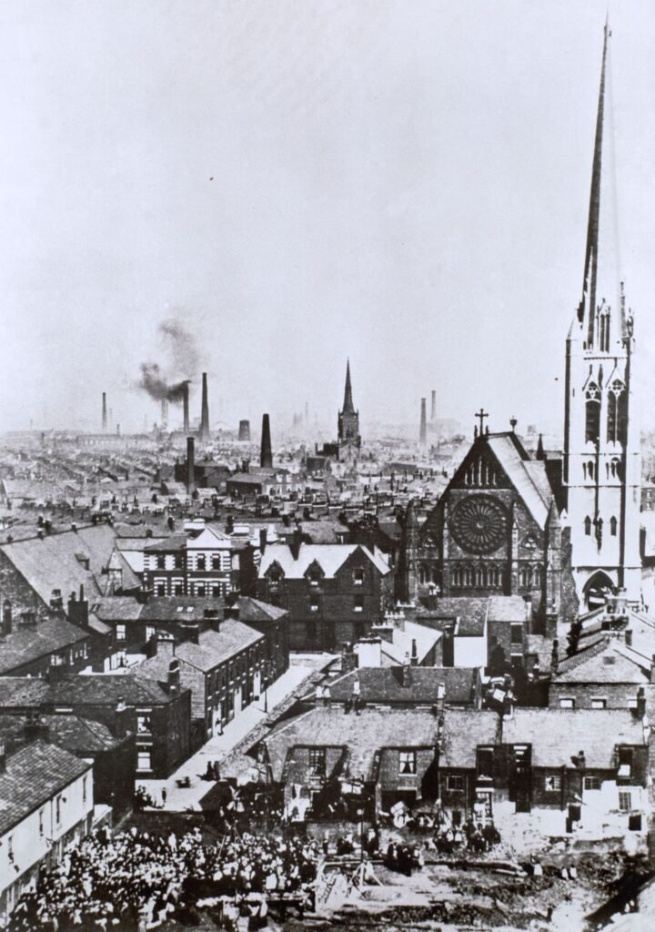 Historical photograph looking over Preston's rooftops and mill chimneys, St Walburge's rose window and spire rising above the terraces, a crowd gathered in the street below.