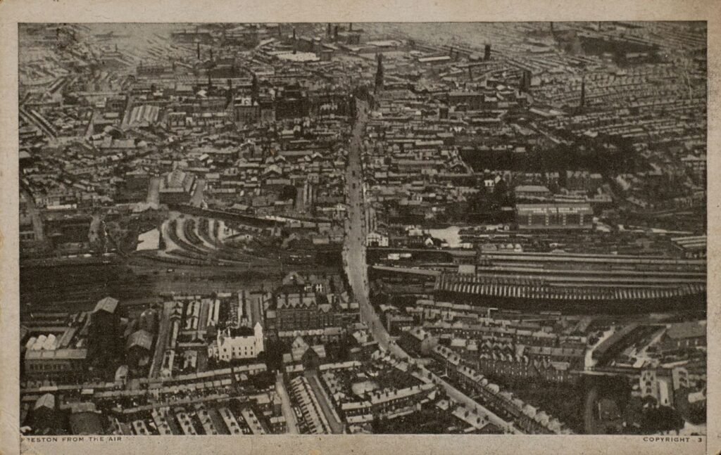 Printed postcard of Preston seen from the air, dense terraced streets and a railway station and sidings running across the middle distance, captioned "Preston from the Air."
