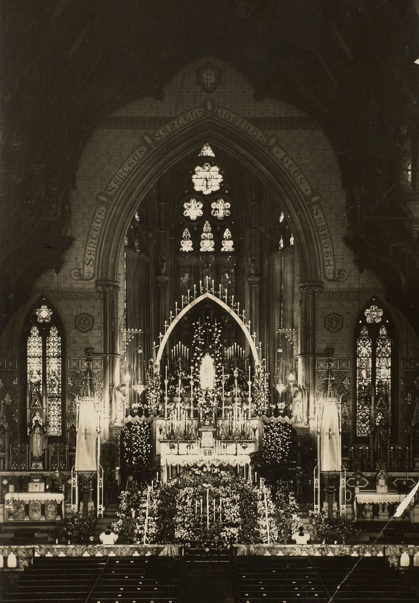 Photograph of St Walburge's sanctuary banked with tiered candles and flowers before the east window, two servers kneeling below.