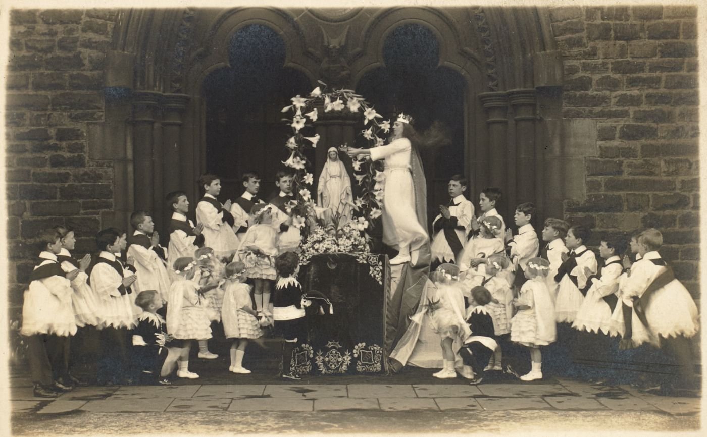 A crowned girl in white reaches to place flowers or a crown at a statue of Our Lady, altar servers and children in white gathered around, at the arched entrance of the church.