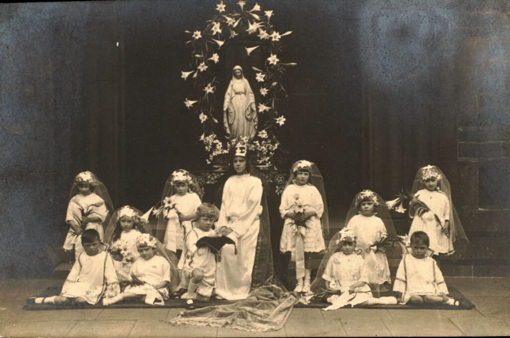 Children in white communion dress and veils, holding lily bouquets, are arranged before a statue of Our Lady wreathed in lilies.