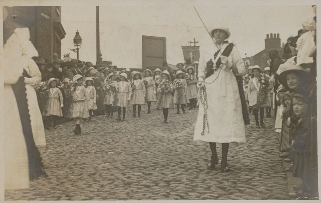 Girls in white dresses and sashes carry flowers along a cobbled street, a woman in white leading with a processional cross behind, crowds watching from the pavement.
