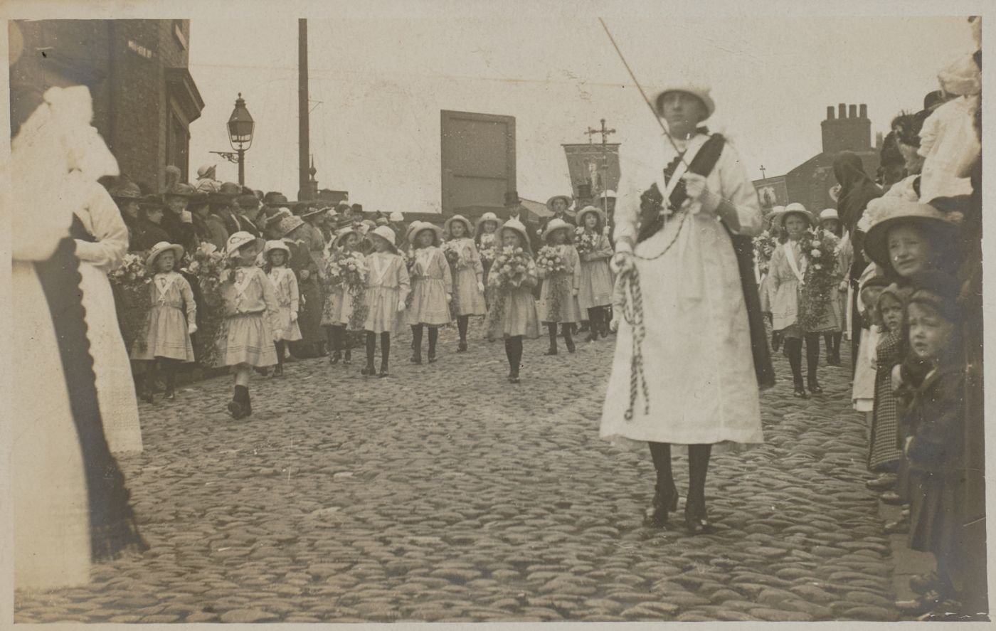 Girls in white dresses and sashes carry flowers along a cobbled street, a woman in white leading with a processional cross behind, crowds watching from the pavement.