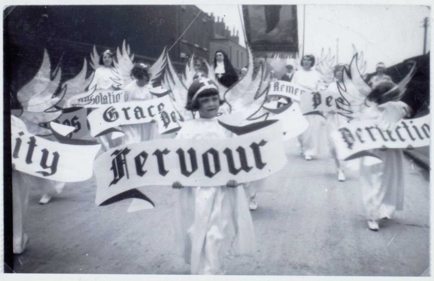 Girls dressed as angels carrying virtue banners including "Fervour" and "Grace," processing down a street.