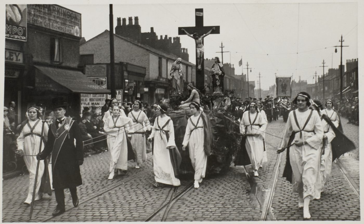 A large Calvary group of the crucified Christ, Our Lady and St John is drawn on a float through a cobbled street, women in white robes and sashes hauling on ropes alongside, crowds watching from the pavement.