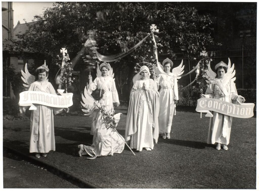 Young women dressed as angels stand around a central figure in a crown and veil with hands joined in prayer, banners reading "Immaculate Conception" held at either end.