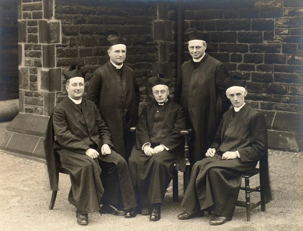 Three clergy sit and two stand for a group photograph outside the stone church buttress.