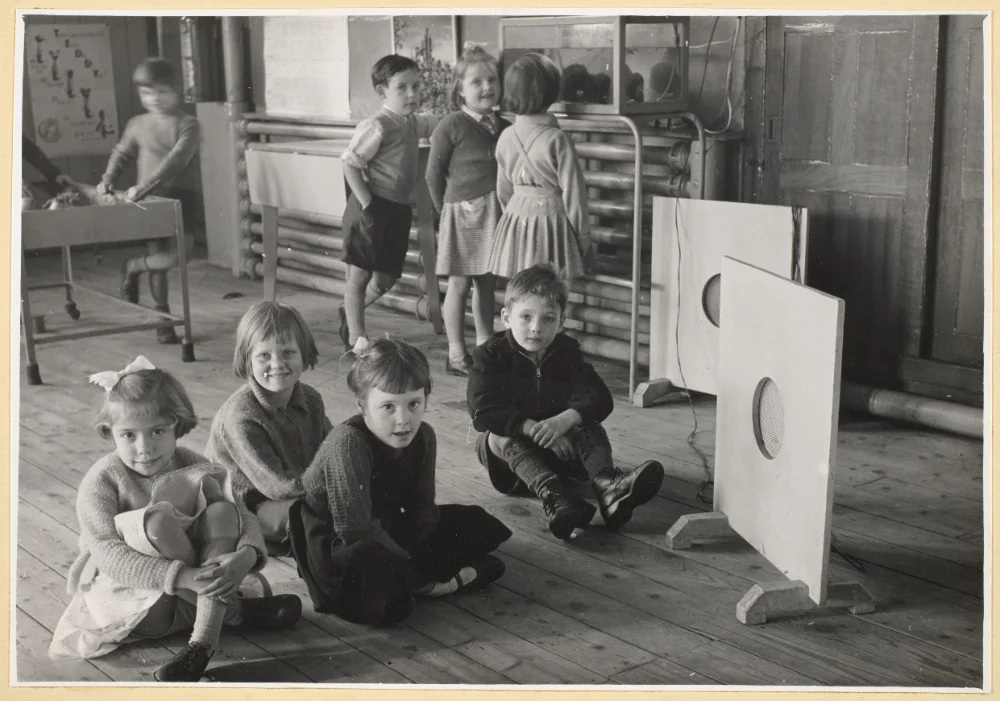 Infant pupils sit and stand around the school hall. 