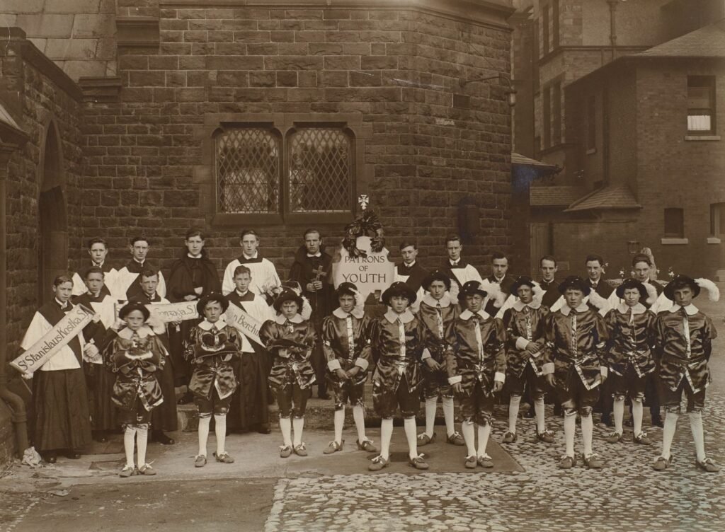 Boys dressed as patron saints of youth stand for a group photograph, altar servers and banners naming individual saints behind, a central banner reading "Patrons of Youth".