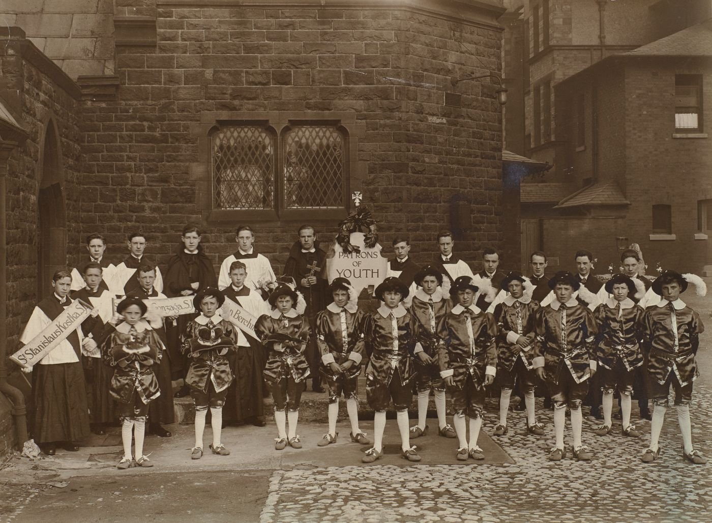 Boys dressed as patron saints of youth stand for a group photograph, altar servers and banners naming individual saints behind, a central banner reading "Patrons of Youth".