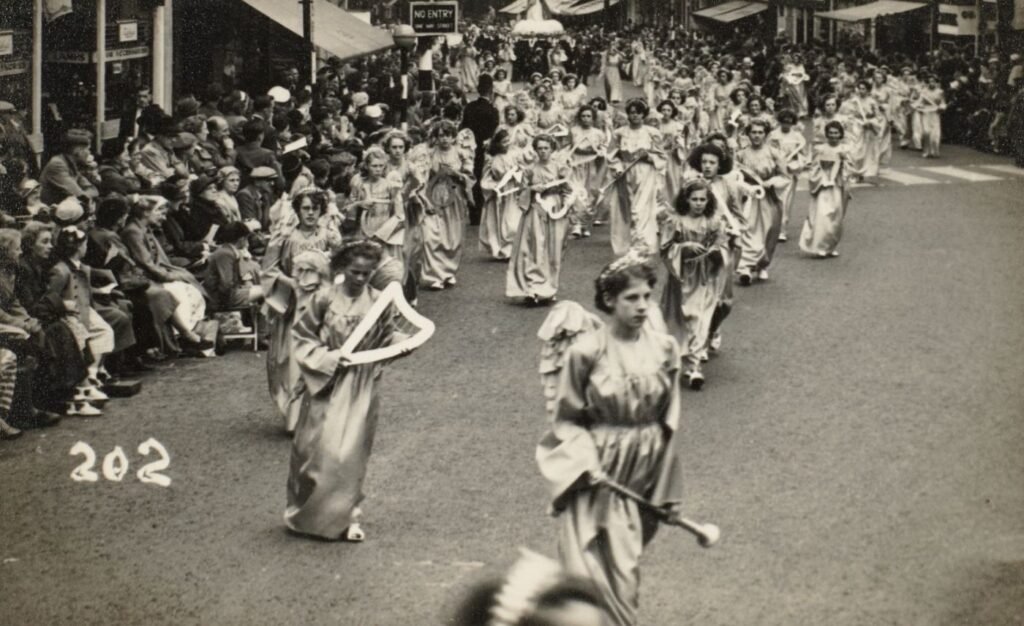 Girls dressed as angels with model harps and trumpets, processing down a Preston street, crowds seated along the pavement.