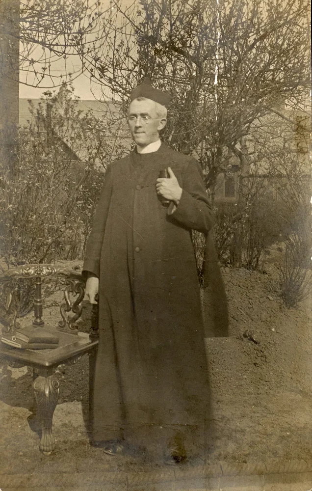 A priest, wearing glasses and a biretta, stands in a garden beside an ornate table holding a book, one hand resting on a chair