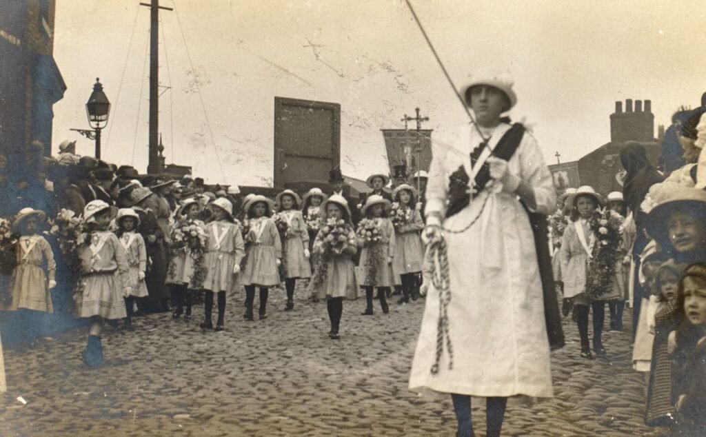 Girls in white dresses and sashes carry flowers along a cobbled Preston street, a woman in white leading with a processional cross behind, crowds watching from the pavement.
