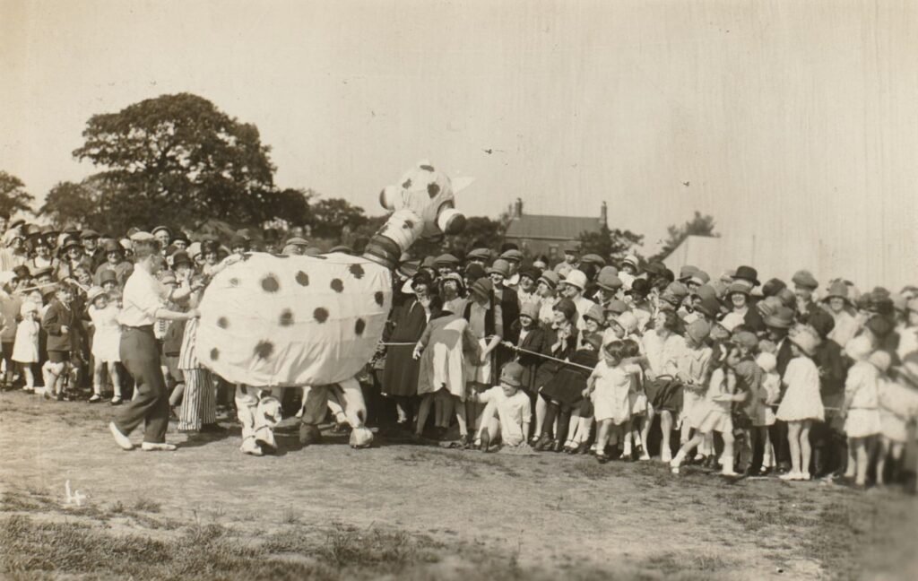 A comic two-person animal costume, spotted and long-necked, is led past a crowd of parishioners and children lining the field.