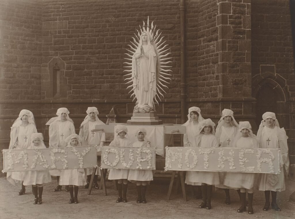 Girls and women in white veils pose beside a statue of Our Lady, holding flower-decorated banners reading "Mary Our Mother."