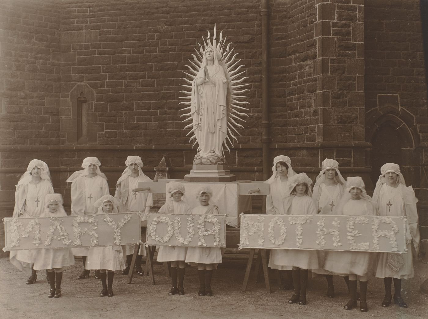 Girls and women in white veils pose beside a statue of Our Lady, holding flower-decorated banners reading "Mary Our Mother."