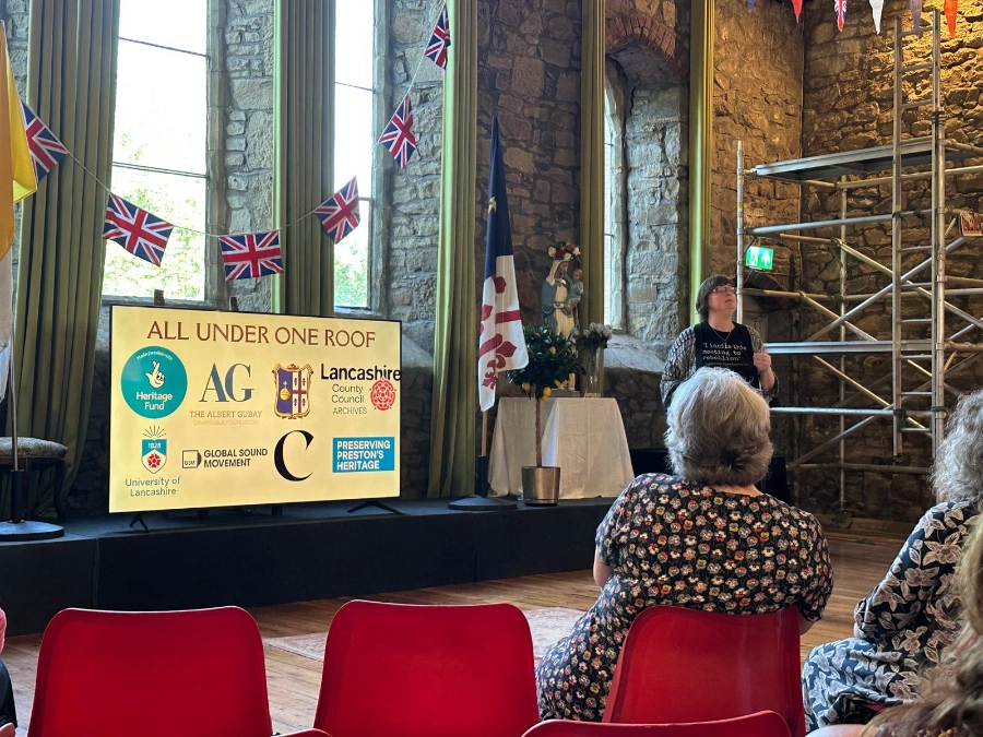 Audience listening to a speaker at the Exhibition launch event, beside a screen reading "All Under One Roof" with partner and funder logos.