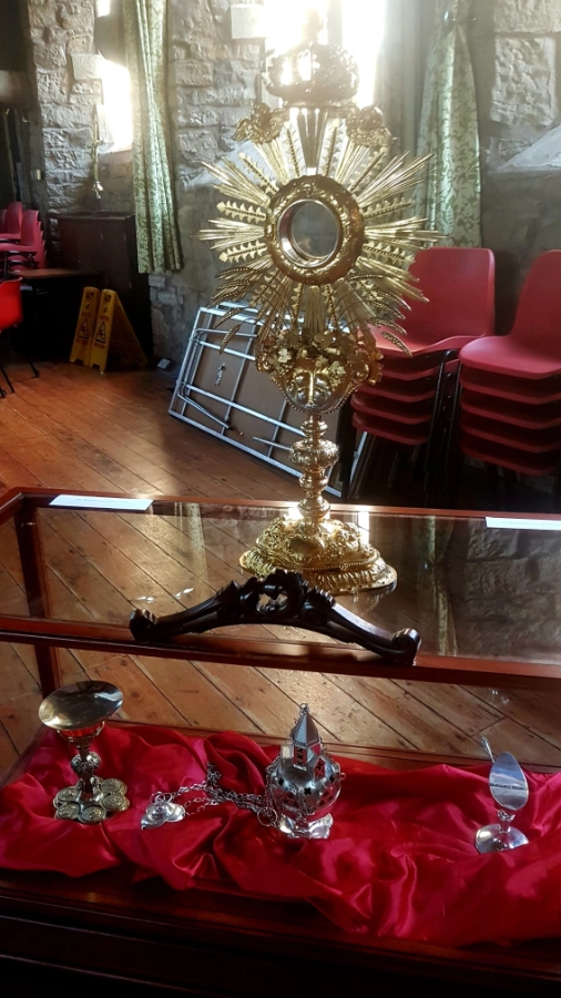 A gold monstrance on a display case holding a chalice, censer and other silver church items on red satin.