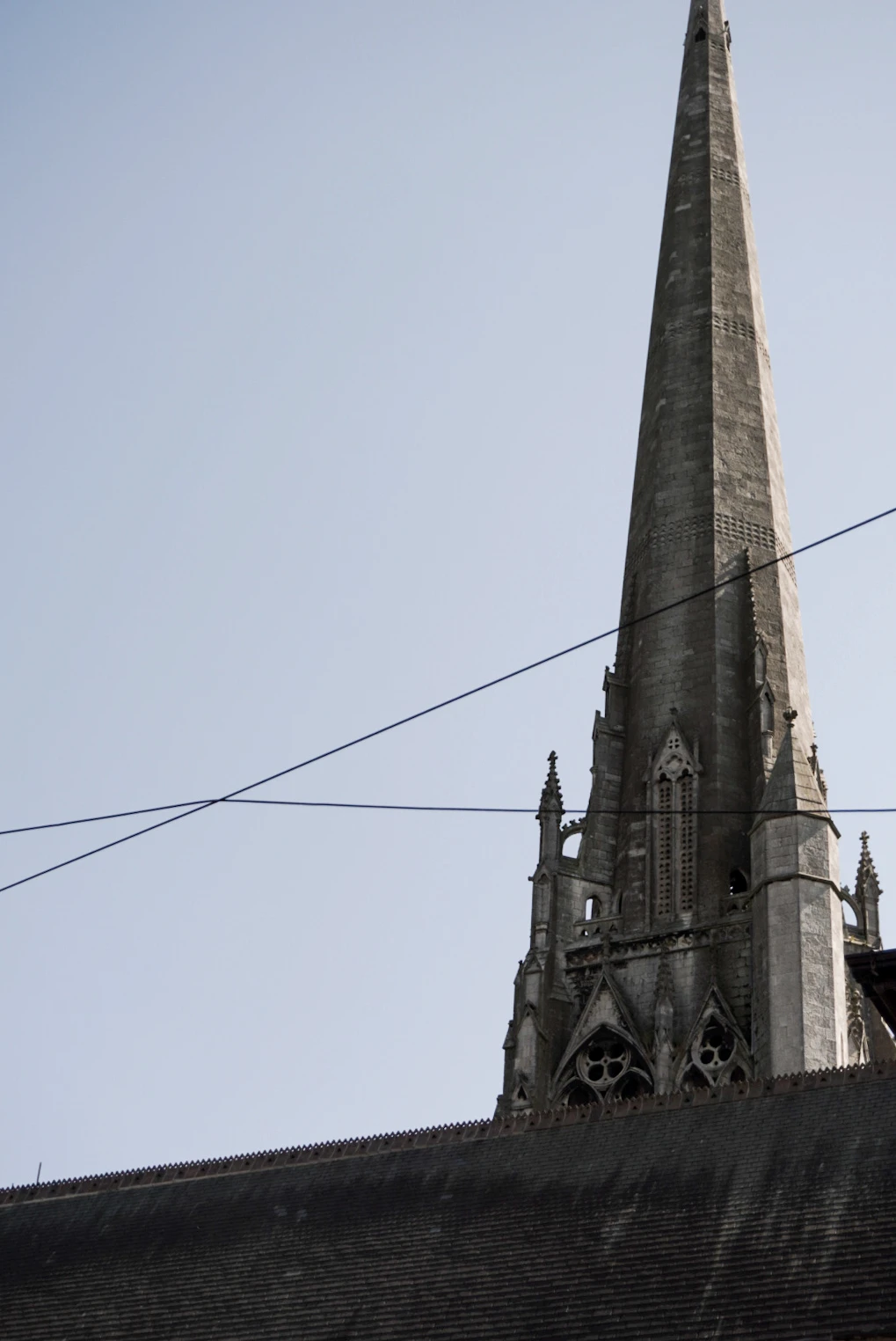 The tall stone spire of St Walburge's Church rising above its dark slate roof against a pale blue sky, with overhead cables crossing the view.