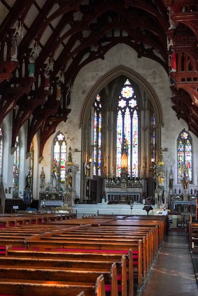 Interior of St Walburge's Church, looking down the nave past rows of wooden pews towards the high altar and a tall stained glass window. Carved timber roof beams arch overhead, with small painted statues set along them.